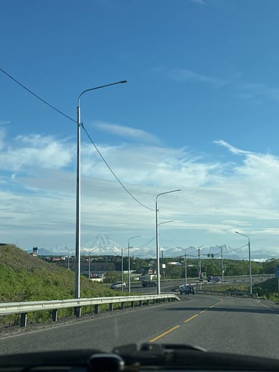 The image shows a view of a road in the city of Svetly, Russia, with a background of snow-capped mountains. The road curves to the right, with a metal guardrail on the left. Several streetlights are visible along the road. In the distance, a town or city can be seen nestled between the road and the mountains. The sky is blue with scattered white clouds. The scene appears to be captured from a vehicle, as indicated by the dashboard visible at the bottom of the frame.