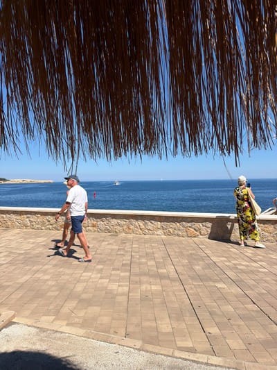 The photograph, taken in Cala Rajada, Spain, features an outdoor scene along a brick-paved walkway bordering the ocean. A thatched roof structure partially obscures the left side of the frame. The clear blue sky meets the deep blue sea, with a small sailboat visible in the distance. On the walkway, a man wearing a white t-shirt and blue shorts is walking. Further down the walkway, a woman in a colorful floral dress and a hat is standing by a short stone wall, seemingly observing something with a cell phone. The walkway is lined with a stone retaining wall. The overall setting suggests a coastal tourist area.