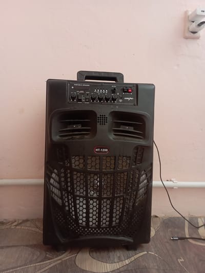 A black portable speaker with the model number HT-1200 is positioned vertically on a patterned floor. The speaker has a handle at the top, a control panel with various buttons and inputs, and a large grille covering the speaker cone. Two white electrical outlets with plugs are mounted on a light pink wall behind the speaker. A black cable is connected to the speaker and runs across the floor. The overall environment appears to be indoors, with the light pink wall and patterned floor suggesting a room. The scene is static and there are no people present. The specific location of Alğa, Kazakhstan is not visually discernible from the image itself, but the description is provided as per the prompt's instruction.