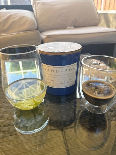 A close-up, high-angle shot shows a glass of espresso and a glass with lime slices on a dark, woven table. A blue scented candle with a wooden lid is positioned between the two glasses. In the blurred background, a white sofa with cushions is visible. The scene is outdoors, possibly on a patio or balcony, suggested by the natural light and outdoor furniture. The overall impression is one of relaxation and leisure. The image does not contain any identifiable landmarks or context that would place it in Colchester, United Kingdom.