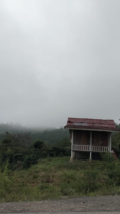 In Bintangpermata, Indonesia, a small, wooden house with a red-tiled roof sits on stilts on a grassy incline. The house features white railings on its porch and a dark-colored door. The surrounding environment is lush with greenery, with trees and vegetation visible on the rolling hills in the background. The sky is overcast, with a thick layer of fog or clouds obscuring the distant landscape, creating a muted and atmospheric scene. A gravel road is visible in the foreground.
