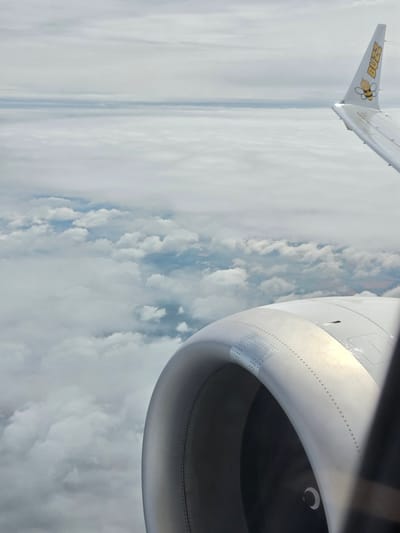 This is an aerial photograph taken from the window of an airplane. The image prominently features the engine nacelle of the aircraft in the foreground, which is metallic and cylindrical in shape. A portion of the airplane's wing is visible on the right side of the frame, featuring the "Buzz" airline logo with a cartoon bee. The background is filled with layers of clouds, creating a soft, hazy atmosphere. The terrain below is obscured by the clouds, but hints of fields and possibly roads can be seen in patches where the cloud cover is thinner. The sky is overcast with a light, diffused light. There are no people or animals visible in the image. The scene depicts a typical view from an airplane during flight, suggesting travel at a high altitude above a cloudy landscape. The location is identified as the city of Orliak, Bulgaria, implying this aerial view is of the airspace above or near that region.