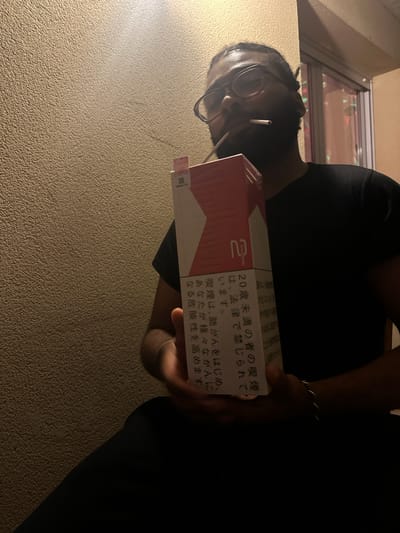 The image depicts a person holding a pack of cigarettes, specifically Marlboro 2's, with Japanese text on the side, including a warning about the dangers of smoking for those under 20. The person, who has dark skin, wears glasses and has a cigarette in their mouth. The photo is taken from a low angle, looking up at the person. The environment appears to be indoors with an off-white wall and a window visible in the background. There is no indication that this image was taken in Montreuil, France.