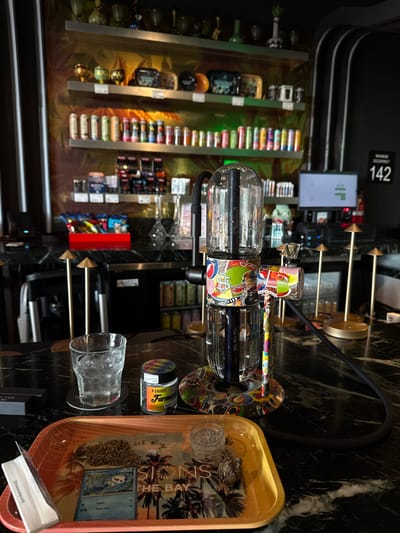 An indoor scene in National City, United States of America, depicts a commercial establishment, likely a cannabis lounge or dispensary, focusing on a dark marble-patterned countertop. In the foreground, a light orange/yellow tray with a palm tree graphic and the text "SIONS THE BAY" holds a pile of ground dried green plant material (consistent with cannabis), a small clear empty glass container, a small amount of unground dried plant material, a Pokémon "Chinchou" trading card, and a small white paper packet labeled "Swarajya milk." To the left of the tray is a clear glass containing ice and residual liquid. Behind the glass, a small clear jar with a black lid is labeled "4 GRAMS Farmers." Dominating the center is a large, elaborate gravity bong or smoking device, featuring clear glass chambers, a black hose, and a base adorned with colorful pop-art style graphics, including dollar signs. It has a bowl attachment on the right side.
In the midground, behind the counter, a red tray contains various packaged snacks. Behind this, shelves display numerous packaged products, some cylindrical and brightly colored. A black structural element runs vertically on the right. A computer monitor displays green bar graphs. A sign on the wall indicates "MAXIMUM OCCUPANCY 142." The background reveals multiple shelves against a warm-toned, patterned wall, displaying a variety of glass items, packaged goods, and cylindrical containers. No individuals are visible. The setup suggests preparation for the consumption of cannabis.