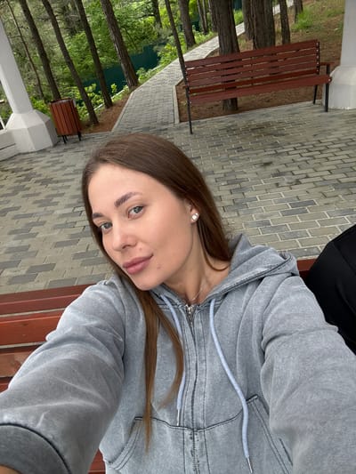 A woman sits on a brown wooden bench in Sosnovy Bor, Russia. The city is visible in the background, with trees and a gray brick walkway near a white building. The woman is wearing a gray hooded jacket. The bench is located in a park or recreation area.