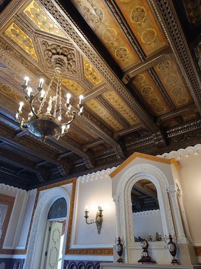 The image shows an ornate, dimly lit interior of a room, likely a historic building or palace, with a heavily decorated wooden ceiling and a large, brass chandelier. The ceiling features intricate carvings and painted panels with geometric and floral motifs in gold, green, and blue. The walls are painted in a neutral tone and feature decorative molding. In the foreground, there is a white, arched mantelpiece with a large mirror reflecting the ceiling and chandelier. Below the mirror, there is a mantel clock and two decorative vases. A wall sconce with two candle-like lights is visible on the right wall. The overall impression is one of opulent and detailed craftsmanship. Based on visual information from other sources, this interior is located in the Nesvizh Castle in Belarus, which is within the administrative region of Краснабярэжскі сельскі Савет.