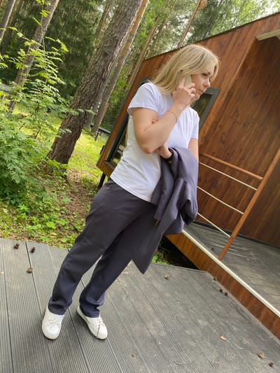 A woman with blonde hair is standing on a wooden deck next to a modern wooden structure, possibly a cabin. She is wearing a white t-shirt, dark grey pants, and white sneakers. She is holding a dark grey jacket over her left arm and talking on a cell phone, holding it to her right ear. The deck is made of dark grey planks and has several pinecones scattered on it. The background shows a dense forest with tall trees and green foliage. The overall impression is a serene, natural setting. The location is not specified as Noviy, Russia, but is a forest or wooded area.