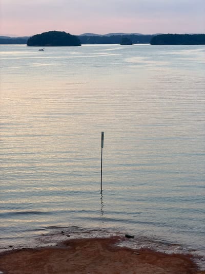 This image presents a serene landscape of a large body of water, identified as being in Sugar Hill, United States, at either dawn or dusk, indicated by the soft, diffused light and pastel sky colors. The top quarter of the frame shows a clear sky with a gradient of pale purplish-pink at the horizon transitioning to a lighter, almost white-blue overhead. Along the distant horizon, several dark, tree-covered islands are visible, silhouetted against the lighter sky, with faint outlines of hills or mountains extending further back.
The expansive water surface, which dominates the midground, exhibits gentle, rhythmic ripples, reflecting the sky's soft hues in muted blues, grays, and subtle warm yellows. A small, white motorboat is present in the mid-distance, positioned to the left of the largest central island. In the foreground, on the right side of the frame, a slender dark pole with a white, rectangular marker attached to its top extends vertically from the water. The immediate shoreline in the bottom portion of the image is composed of reddish-brown earth, where small waves are gently breaking. A dark, elongated object, resembling a shoe or piece of debris, is partially submerged at the water's edge on the right, near several smaller stones. No people are visible in the scene.