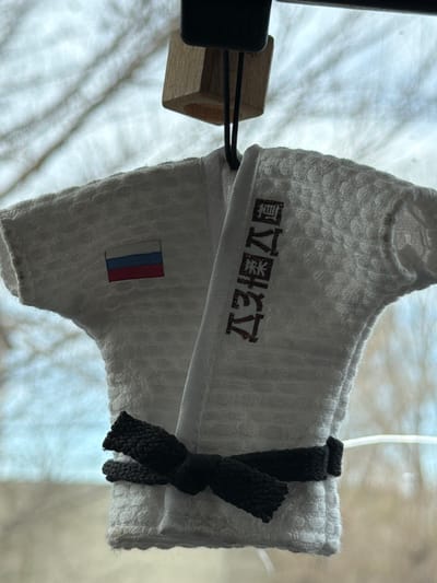 The image shows a white, textured towel with Japanese characters printed on it, along with a small Russian flag patch. The towel is suspended, possibly hanging, and secured with a black woven tie. The setting is difficult to discern, but it appears to be indoors with a white or light-colored background, possibly a sheet or curtain. The scene is not clearly identifiable as being specifically in Расцвет, Russia.