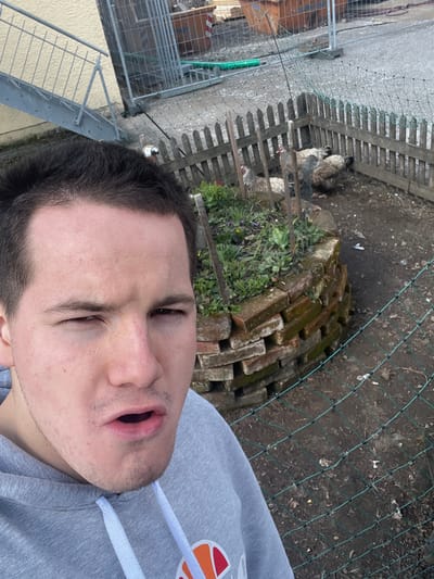 The image shows a man with dark hair and a grey hoodie in an outdoor setting. The man's face is prominent in the foreground, with his mouth slightly open. Behind him, there is a fence and what appears to be a chicken coop with chickens. There are also bricks stacked near the fence. The environment includes dirt and vegetation. There is a metal structure to the left side of the frame. The location is identified as Benningen, Germany.
