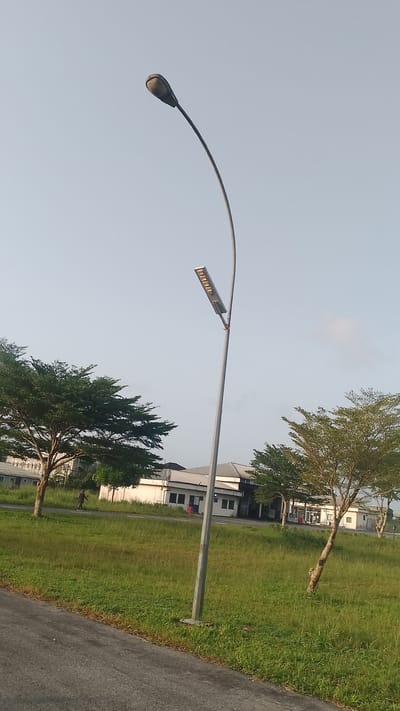 The image depicts an outdoor scene in Effurun, Nigeria. A tall, curved street lamp is the central focus, with two different lamp types visible on the same pole. An older-style, opaque lamp is positioned at the top of the curve, while a newer, rectangular LED panel is mounted lower on the pole.
The background features a low, white building with a metal roof, typical of commercial or industrial structures. Several medium-sized trees with green foliage are scattered around the lawn in front of the building. A single person is visible walking in the vicinity. The sky is a clear, light blue. The immediate foreground consists of a grassy area, with an asphalt road running along the bottom edge of the frame.