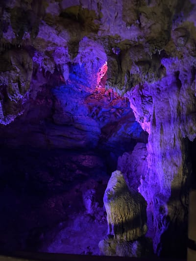 The image depicts the interior of a cave, likely a tourist attraction, illuminated by artificial colored lights. The cave walls are a complex formation of rock, with stalactites hanging from the ceiling and stalagmites rising from the floor. The dominant colors are deep purple and vibrant blue, with some areas of pink and yellow light highlighting specific rock formations. The texture of the rock is rough and varied, with smooth, layered surfaces and jagged, crystalline structures. The lighting creates dramatic shadows and highlights, emphasizing the geological features of the cave. It is impossible to determine the specific location within Sanguansi, China from the image alone, as no identifying landmarks or signage are visible. There are no people or animals present in the scene. The overall impression is one of natural beauty enhanced by artificial illumination.
