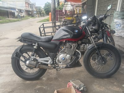 A black and red motorcycle with a BMW logo on the engine block is parked on a dirt and concrete surface in front of a shop with a corrugated metal roof. The motorcycle has a custom-built metal rack attached to the rear. To the right of the motorcycle is a large, white, woven bag with text that appears to read "178" and some other Indonesian words. In the foreground, on the ground, are a disassembled engine part and a cardboard box. In the background, a street lined with buildings and palm trees can be seen. This scene is located in Rambot, Indonesia. There are no people clearly visible in the image.