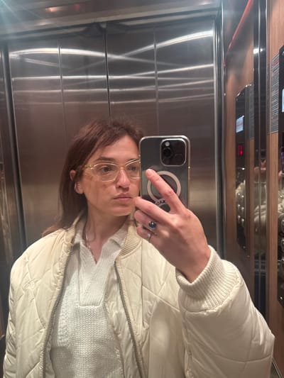 The image captures a mirror selfie taken by a person in an elevator. The photo is rotated 90 degrees counter-clockwise, making the elevator walls appear horizontal instead of vertical.
In the foreground, a person is holding a smartphone to capture their reflection. The person has medium brown, shoulder-length hair and is wearing light-colored, possibly clear or yellowish, square-framed glasses. Their face is partially visible, showing their forehead, eyes, nose, and chin. They have a neutral expression.
The person is dressed in a light-colored, possibly cream or off-white, quilted jacket with ribbed cuffs and collar. Beneath the jacket, a white or cream knitted sweater with a V-neck is visible. On their right hand, which is holding the phone, two rings are worn on the ring finger: a prominent dark blue or black stone ring with a silver-colored band, and a thinner silver-colored ring next to it.
The smartphone appears to be an iPhone, identifiable by its triple camera array on the back. It is encased in a clear or translucent protective case, through which a white MagSafe ring is visible. The text "MAX" is faintly visible on the case near the MagSafe ring. The back of the phone is reflecting light, and the screen is not visible.
The background clearly indicates an elevator interior. The walls are made of reflective, brushed stainless steel panels, creating vertical lines and reflections of light. At the top of the image (which would be the top of the elevator in a correct orientation), an elevator control panel is mounted on a wooden-look trim. A red digital display on the panel shows a number (possibly '2') and an arrow pointing to the right (which would be 'up' in a correctly oriented panel). Other smaller, illegible text and button outlines are also present on the control panel. The overall lighting seems bright, reflecting off the metallic surfaces of the elevator and the phone.