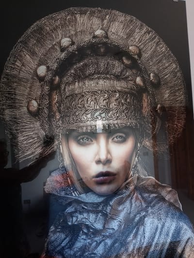 The media file is a photograph featuring a close-up portrait of a woman adorned in an elaborate, ornate headdress. The headdress is large and circular, with intricate detailing, appearing to be made of woven materials and decorated with small, round, pearl-like objects. The woman has striking blue eyes and is wearing dramatic makeup, including dark lipstick. She is also wearing a shimmering, metallic-like garment that drapes around her shoulders and neck, and a large, decorative earring. The lighting suggests a studio setting, highlighting the textures and details of her headdress and attire. Behind her, a shadowy figure can be seen, possibly a photographer or assistant, with their arm extended, suggesting the photograph was taken during a photoshoot. The overall mood is artistic and theatrical, with a focus on the woman's regal and striking appearance. No text is visible in the image. The location context provided is Rivodutri, Italy.