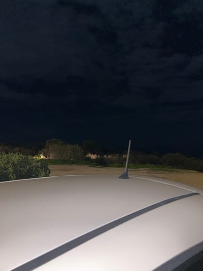 An outdoor scene at night, taken from an elevated perspective, possibly a balcony or boat deck, in Anatoliki Attiki, Greece. The right side of the image is dominated by a smooth, white, curved structure with a distinct gray line, which frames the view. A slender, dark-colored pole or antenna extends from this white structure towards the center of the frame.

In the middle ground, a strip of land with vegetation, including bushes and trees, is visible. Parts of this foliage are subtly illuminated by artificial lights, creating soft glows against the darkness. To the left, a large, dark expanse occupies the rest of the frame, showing some texture that suggests either clouds in a night sky or the surface of a body of water. The overall atmosphere is dark and tranquil, indicative of nighttime. No people or specific activities are visible, and there is no discernible text.