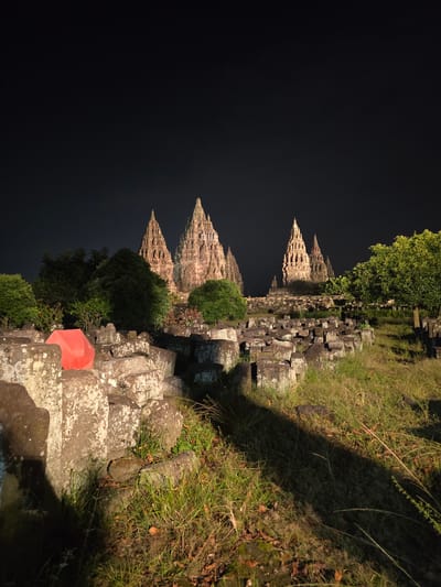 The image depicts the Prambanan Temple compound in Bokoharjo, Indonesia at night. The spires of the main temples are visible against a dark sky. Stone structures and a bright red parasol form a pathway into the foreground. Vegetation, including grass and trees, flanks the pathway. The image has a slight tilt, rotating the right side forward.