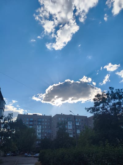 This is a daytime photograph taken outdoors, likely in a residential area of Alchevsk, Ukraine. The scene is dominated by a bright blue sky with scattered white clouds. A large, dark cloud is positioned in the center, with sunlight streaming through it, creating visible crepuscular rays.

In the foreground, the top of a multi-story apartment building is visible, with multiple windows and balconies. Dark green trees partially obscure the building and line the bottom of the frame. Below the trees, a paved area appears to be a parking lot or street, with several parked cars visible. A single streetlight stands near the apartment building. The overall impression is that of an ordinary day in a city, with a focus on the interplay of light and shadow from the sky. The time of day appears to be late afternoon or early evening, given the angle of the sunbeams. No people are clearly visible in the image. There is no visible text in the image.