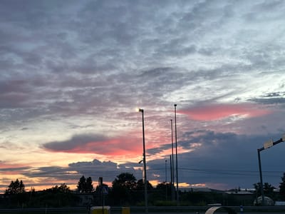 The image depicts a sunset over Laagri alevik, Estonia. The sky is filled with dramatic clouds tinged with pink and orange hues, contrasting with darker, stormy gray clouds. Several lampposts stand tall in the foreground, with one illuminated lamp. In the middle ground, a line of trees separates the sky from buildings and structures in the distance. A road with traffic lights and road signs is visible at the bottom of the frame. The overall scene captures the transitional light of dusk in a semi-urban environment.
