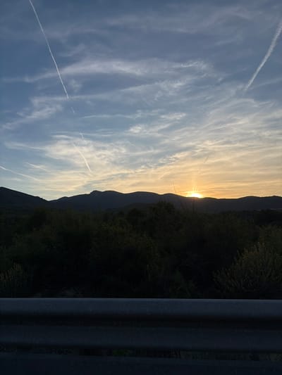 The image shows a sunset in Urbanització El Totxar, Spain. The sun is setting behind a mountain range, and the sky is filled with streaky clouds and contrails from airplanes. To the right of the mountains is a dark area of greenery, which could be trees or bushes. On the far right of the image is a gray, metallic structure, possibly a guardrail.