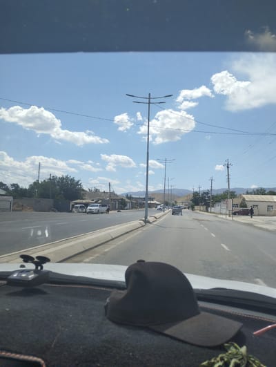 A frontal, ground-level, eye-level, medium shot depicts a sunny day on a paved road in the city of Forish QFY, Uzbekistan. The road is lined with trees, buildings, and utility poles. In the foreground, a black baseball cap sits on the dashboard of a vehicle, partially obscuring the view of the road ahead. Several cars, including white vans and sedans, are visible on the road, as are a few pedestrians. The sky is a clear blue with scattered white clouds. The overall scene conveys a sense of everyday life in a semi-urban environment.