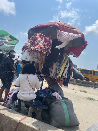 The image displays an outdoor market scene under a bright, partly cloudy sky in Aiyetoto-Asogun, Nigeria. In the foreground, a person with dark, curly hair, wearing a white t-shirt and dark pants, sits on a wooden bench, facing away from the camera, organizing or examining a pile of clothes. A dark shoulder bag is visible across their back. Adjacent to them, two large, dark grey woven sacks with a vertical green stripe rest on the ground. A large, predominantly red and maroon market umbrella, heavily laden with various hanging clothes, including patterned shirts, dresses, and blouses, stands centrally. Another green and white market umbrella is partially visible on the far left. To the left of the seated individual, a person wearing a dark, patterned shirt, dark shorts, and a light-colored visor stands, appearing to sift through clothing items. The ground is unpaved and dusty, transitioning to a raised concrete curb in the immediate foreground and a road in the background where a yellow commercial bus and a dark passenger car are visible, along with additional indistinct figures.