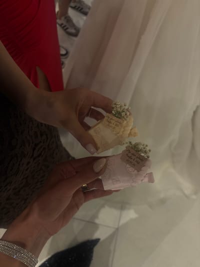The image shows two hands holding small house-shaped objects, likely wedding favors. One house is cream-colored, and the other is pink. Each house has a small tag with the text "Մեր սիրո տնակը, Դավիթ և Անի, 03.05.26", which translates to "Our Love House, Davit and Ani, 03.05.26", indicating the names of a couple and a date. Both houses are adorned with small white flowers. The person on the left is wearing a red dress, and the person on the right is wearing a diamond bracelet. The location, Masis, Armenia, cannot be confirmed from the image alone.