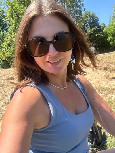 A woman with brown hair and sunglasses is pictured outdoors, seated on dry grass. She is wearing a light blue sleeveless top and a silver necklace. Her right arm is visible, and she appears to be holding a phone, which is reflected in her sunglasses. Behind her, trees and a clear blue sky are visible, suggesting a warm, sunny day. The background appears to be a rural or park setting, possibly in the Coteaux du Lizon region of France.