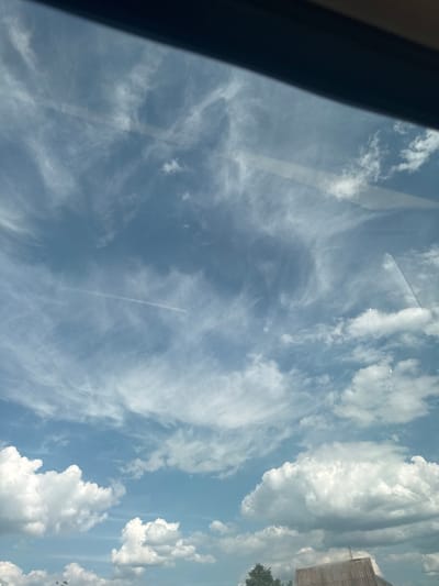 The image displays a daytime view of the sky over Markivka, Ukraine, captured through a reflective surface, likely a window or windshield, indicated by subtle glare and reflections. The upper and middle portions of the sky are dominated by light blue hues, traversed by widespread, wispy cirrus clouds forming intricate, feathery patterns. A distinct, linear contrail, indicative of an aircraft's passage, extends horizontally across the central-upper sky. Towards the bottom, several bright white, puffy cumulus clouds are present. The lower-most section of the image reveals elements of the ground, including a dark, sloped, corrugated roof structure on the right and a portion of dark green foliage, possibly from a coniferous tree, on the left. A dark, solid, diagonal obstruction is visible across the top of the frame. No individuals, specific actions, or direct interactions are discernible within the frame.