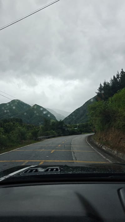 A driver's perspective photograph depicts a winding road in a mountainous region, presumably near Bình Lư, Vietnam based on the prompt. The asphalt road features yellow divider lines and appears to be damp or wet, possibly due to rain. The sky is overcast with heavy clouds. Green, tree-covered mountains rise on either side of the road, receding into the distance with a visible layer of mist or haze. The foreground shows the dashboard of the vehicle from which the photograph was taken, with water droplets on the windshield.