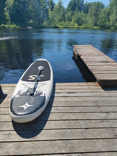 A stand-up paddleboard with a paddle lying on it rests on a wooden dock. The paddleboard is white with black and grey accents, including a logo that resembles a stylized bird and the letters "ST". The dock is made of weathered wooden planks and extends into a dark, calm body of water. Beyond the water, a dense line of green trees forms the background under a clear blue sky. The scene is bathed in bright sunlight, suggesting it is a sunny day, likely midday or afternoon. There are no people visible in the image, and the overall atmosphere is peaceful and serene. The location is Zashchita, Russia.