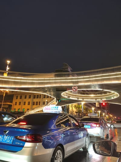 This is a night-time, medium-shot, eye-level photograph taken in Jixi, China, likely on a street with a pedestrian overpass. The scene depicts a traffic jam with several cars, including taxis, stopped on a wet road.

In the foreground, the rear of a blue and silver taxi is visible. Its license plate reads "G TP327". The taxi has a prominent illuminated "TAXI" sign on its roof, with the number "12328" displayed. The rear brake lights are illuminated red. To the left, the side of another vehicle, likely another taxi, is partially visible. A side-view mirror, reflecting a person's hand, is in the lower right corner of the frame.

Behind the first taxi, a purple and silver taxi is also stopped, with its license plate reading "G TO171". Further back, other vehicles and the red light of a traffic signal are visible.

The environment includes a multi-level pedestrian overpass illuminated with a continuous line of bright white lights, creating a glowing effect. The overpass has a metallic railing. In the background, buildings with lit windows are visible, suggesting an urban setting. The dark night sky is above. The overall impression is of a busy city street at night, possibly after rain due to the wet road surface.