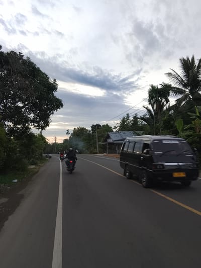 The image depicts a road scene in Meunasah Kumbang, Indonesia, under a cloudy sky at dusk. A black minivan is driving on the right side of the paved road, which is marked with a yellow line. A person on a motorcycle is in front of the minivan, also traveling on the road. The left side of the road is bordered by lush green foliage and trees, including a large tree on the far left. On the right side of the road, there are palm trees and houses. Electrical poles and wires are visible in the background. The overall impression is that of a rural or suburban street in a tropical region.