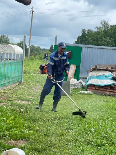 A male individual operates a gas-powered string trimmer in a grassy outdoor area in the city of Bormist, Russia. The man wears a blue and light blue work overall, black rubber boots, and a dark blue baseball cap with a white "NY" logo. He holds the trimmer's handles, with the orange and black engine unit visible on his right side, actively cutting through the green grass and dandelions in the foreground. In the midground to the left, a light green mesh fence precedes a white hoop-style greenhouse and a tall utility pole. To the right, a green corrugated metal shed with a small reddish-brown mailbox-like object is visible, next to a larger grey corrugated metal structure. Between these sheds, a pile of red bricks is partially covered by a white and blue tarp, topped with wooden planks. A second individual, appearing female with light hair, is observed in the deeper midground, bent over with a long-handled tool in an overgrown area. The background features a dense line of green trees under an overcast, light grey sky with visible utility lines. The scene depicts a semi-rural or rural environment.