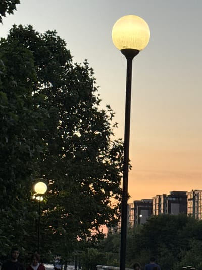 The image shows an outdoor scene at dusk, likely in Otf Kaktugay, Russia. In the foreground, a tall, dark lamppost with a glowing spherical light fixture stands prominently on the right side of the frame. To the left, dense green foliage from trees obscures a portion of the scene. Another, smaller lamppost with a lit globe is visible within the trees towards the lower left. In the background, several multi-story buildings with illuminated windows are visible against a gradient sky transitioning from pale blue to orange. The lower portion of the image shows the tops of several people, some walking, but their features are indistinct due to the low light and distance. The overall atmosphere suggests an urban park or walkway area at the end of the day.