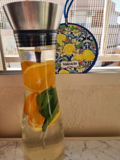 The image displays a clear glass pitcher containing water infused with sliced oranges and fresh mint leaves, topped with a metallic lid. To the left, a circular decorative object, possibly a fan, features a blue braided edge. Its design showcases yellow lemons, white lemon blossoms, and green leaves against a blue and white patterned background, evocative of traditional ceramic artistry. The word "Valencia" is inscribed on a white label within this design.

The background reveals a brightly lit outdoor scene through a window or balcony opening, partially obscured by a white square-grid screen. Beyond the screen, parts of a building's exterior are visible. To the right of the pitcher, there's a section of a textured, light-colored wall, and an adjacent piece of white marble with grey veining. The bright lighting suggests it is daytime, likely a sunny morning or afternoon. The "Valencia" text on the decorative item, combined with the Mediterranean-style lemon motif, strongly hints at the given location context of Xàtiva, Spain, which is near Valencia. No people or specific activities are depicted; the scene primarily focuses on the infused drink and its immediate surroundings.