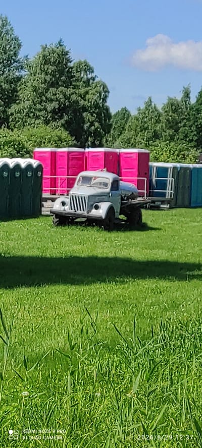 A vintage light gray truck is parked on a grassy field in Võru linn, Estonia. The truck has a distinct vertical grille and round headlights. In the background, rows of portable toilets are visible, with green ones on the left and bright pink ones in the center, followed by teal and dark blue ones on the right. Tall green trees form a backdrop behind the portable toilets. The sky is clear with a few white clouds. The foreground consists of a lush green lawn.