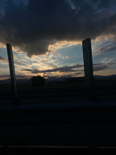 The image captures a sunset over a rural landscape, potentially near Oiejdea, Romania. In the foreground, two dark, vertical metal posts, possibly part of a guardrail or fence, frame the view. Beyond them, a dark road or highway is visible, with a low barrier running along its edge. The middle ground features a flat, open field with a solitary, dark tree in the center. In the distance, rolling hills and mountains form a dark silhouette against the vibrant sky. The sky itself is a dramatic display of sunset colors, with dark, heavy clouds dominating the upper portion, transitioning to lighter, wispy clouds illuminated by the setting sun, which is partially visible and peeking through the clouds. Some structures, possibly buildings or agricultural facilities, are faintly discernible on the horizon. The overall scene is characterized by low light conditions due to the time of day.