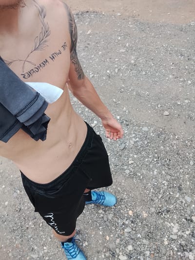 A shirtless man is depicted from mid-thigh up, standing on a ground covered with small grey and white gravel, characteristic of a dirt road or trail in Karnalovo, Bulgaria. He is wearing black athletic shorts and blue athletic shoes with laces. A dark grey shirt, possibly a towel, is draped over his left shoulder and arm, revealing a white tag underneath.
The man has a fit physique with visible abdominal muscles and veins in his right arm. Several tattoos are prominent: on his left chest, there is a design of a feather-like branch. Below this, text is tattooed in a stylized, reversed font, reading "DIE MITW MEMORIES" (likely intended as "DIE WITH MEMORIES"). A sleeve tattoo, appearing to be a tree or nature-themed design, is partially visible on his right arm and shoulder.
The black shorts feature white lettering on the left leg, partially obscured, but resembling "Just" (possibly part of a brand slogan). The blue shoes have visible brand elements on the tongue. The lighting suggests it is daytime, and the ground appears dry. The man is looking downwards.