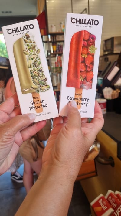In Litochoro, Greece, two hands hold up ice cream packaging. The packaging features the brand "CHILLATO" with "BORN IN GREECE" below it. The left package displays an image of pistachio nuts and a pale green ice cream bar, labeled "130 kcal" and "Real Sicilian Pistachio." The right package shows whole strawberries and raspberries and a red ice cream bar, labeled "Real Strawberry." The background appears to be an indoor setting, possibly a shop, with shelves stocked with various items, and blurred figures of people in the distance.