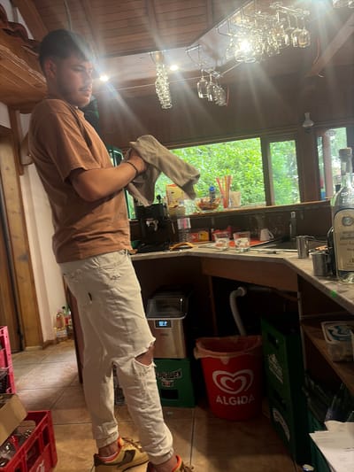 The image depicts a young man standing in what appears to be a bar or restaurant. He is wearing a brown t-shirt and light-colored pants with ripped knees. He is holding a beige cloth in his left hand. The man is looking upwards and slightly to his right with a relaxed expression.

The setting is rustic, with wooden paneling on the walls and ceiling. There are several rows of glasses hanging from a rack on the left side of the image. A counter area is visible in the background, with bottles of alcohol, a coffee machine, and a trash can with an "ALGIDA" logo on it. There are also green crates stacked behind the counter, suggesting a storage area for beverages. Through a window behind the counter, lush green foliage is visible, indicating it might be daytime and possibly a pleasant weather day.

Some visible text includes "ozzo di plomo" on a bottle and "ALGIDA" on the trash can. The overall atmosphere seems casual and informal, perhaps during a quiet period in the establishment.