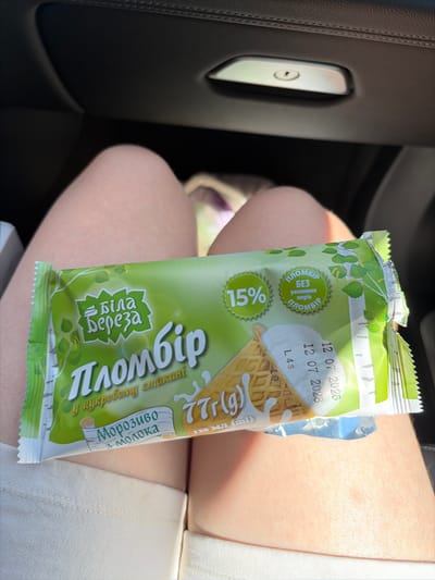 In a car, a person's legs are visible, holding an ice cream cone wrapper. The wrapper is green and features the brand name "Біла Береза" (White Birch) and the product name "Пломбір" (Plombir ice cream). Text on the wrapper also indicates "15%" and "БЕЗ РОСЛИННИХ ЖИРІВ" (Without vegetable fats). A "77г" (77g) weight indication and a date of "12 07 2026" are also present. The image appears to be taken inside a car in the city of Holercani, Moldova.