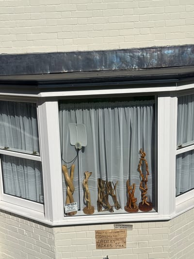 The image displays a cream-painted brick building in Salisbury, United Kingdom, featuring a white UPVC bay window. The bay window comprises at least three sections, with white sheer curtains visible behind all panes. Inside the central window, five natural wood or driftwood sculptures are arranged vertically on the sill; the leftmost sculpture includes a small white "FOR SALE" sign with red text. A small, flat, white rectangular device, possibly an antenna, is attached to the upper interior of the central window pane with a black wire. Below the central window, affixed to the brickwork, is a handwritten cardboard sign stating "MORE BIKES IN STOCK" (partially visible), "ADULT BIKES WANTED," phone number "07856954310," and "LADIES BIKES IN STOCK." Above the bay window, a weathered, dark grey lead flashing or roof element is present. The scene is illuminated by direct sunlight. No individuals are visible.