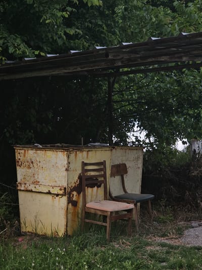 An outdoor scene in Dobri Dol, Bulgaria, shows two dilapidated metal boxes stacked on top of each other, heavily rusted and with peeling yellow paint. Two old wooden chairs, one with a green seat and the other with a pink seat, sit on top of the boxes. Lush green vegetation and grass surround the boxes on one side, while a metal structure stands on the opposite side, with dense trees and foliage filling the background.
