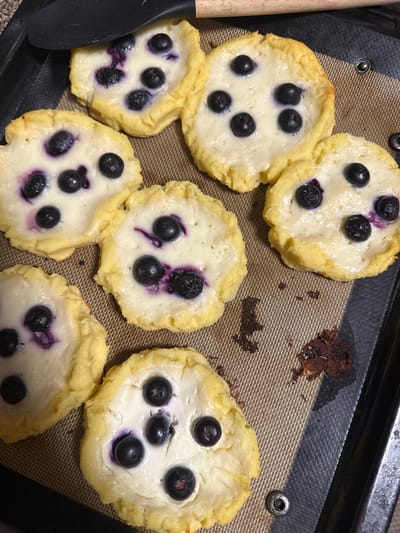 The image depicts a baking tray with seven blueberry and cream cheese pastries. The pastries are irregularly shaped with a pale yellow crust and a white filling dotted with blueberries, some of which have released purple juice into the filling. To the left of the pastries is a black spatula. A few crumbs of what appears to be burnt pastry are visible on the right side of the tray. The scene takes place indoors. There is no evidence to suggest that the image was taken in Vorotynsk, Russia.