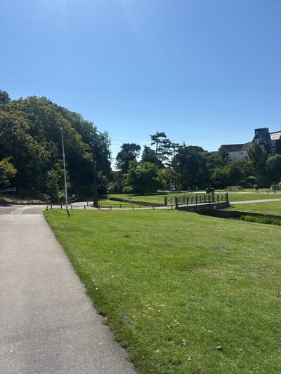 The image depicts a sunny day in a park, likely within Bournemouth, United Kingdom, given the architectural style of the buildings visible in the background. In the foreground, a paved pathway curves to the left, bordered by a well-maintained green lawn. Further into the park, a decorative metal bridge spans a narrow waterway. Lush trees and shrubs form a dense backdrop, with a few buildings peeking through the foliage. A lamppost stands near the pathway, and a bench is occupied by a person sitting with their back to the camera. Several other individuals are seen walking in the distance, enjoying the park. The sky is a clear, bright blue, suggesting fair weather.