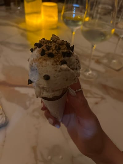 The image depicts an indoor, low-light scene in El Tezal, Mexico, centered on a dessert item. A right hand, with manicured nails painted white and wearing a thin, silver-colored ring on the fourth finger, holds an ice cream cone. The ice cream consists of at least two scoops of a light-colored, possibly white or cream-colored, frozen dessert, topped with dark chocolate chips and light brown, granular crumbs, consistent with crushed graham crackers or similar. Minor melting is visible on the ice cream and the white paper wrapper around the cone. The background, out of focus, reveals a light-colored table surface, possibly marble or stone, reflecting ambient light. Blurred elements include two stemware glasses containing a clear or light-colored liquid, likely wine, and two luminous, cylindrical objects, characteristic of candles or lanterns, casting warm orange light onto the table.
