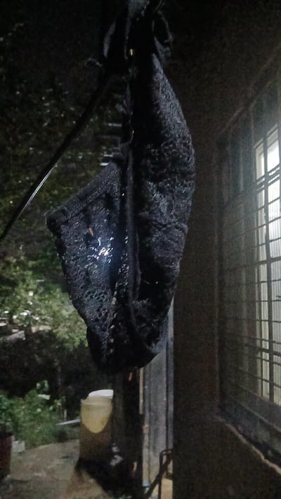An overhead shot captures a dark, lace-like textile draped over a support structure. Rain or moisture is visible on the fabric's surface, reflecting light. Above the textile is a partially visible latticed structure, possibly a roof or covering. The background shows blurred foliage and what appears to be a light source, suggesting an outdoor environment. Based on the provided location, Putucual, Venezuela, this scene may depict an outdoor area within the city, possibly a patio, balcony, or similar space, where the textile is hanging to dry or is being stored.