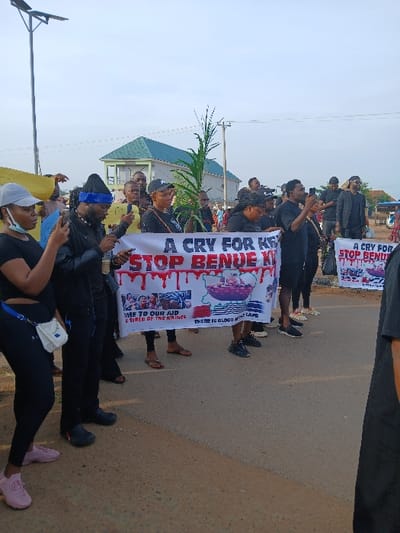 A group of people are gathered on a road, holding banners with the phrases "A CRY FOR BENUE" and "STOP BENUE KILLINGS". The banners depict images of what appear to be blood droplets and a map of Benue State. Most of the people in the group are wearing black clothing. One person is holding palm fronds. Some individuals are raising their hands. The scene appears to be a demonstration or protest.