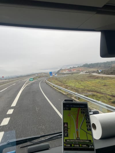The image depicts a forward-facing view from inside a vehicle, likely a truck, traveling on a multi-lane highway (A-24) under an overcast or foggy sky. The vehicle is located on the A-24 highway near Chaves, Portugal, approaching the border with Spain, heading towards Verín, as indicated by the visible navigation system.
The highway, appearing wet, curves gently to the right. White lane markings and dashed lines are clearly visible on the asphalt. To the right of the main highway, an exit ramp leads towards a service area situated on a higher elevation. This service area features several buildings with orange roofs and multiple red and white pylons or flags. The surrounding landscape consists of rolling hills covered in sparse green and brown vegetation, with distant hills partially obscured by fog. Road signs are present on the right side of the highway; one blue sign displays "A-24" and an arrow pointing right, while another further back appears to be a directional sign. A green highway sign with an arrow is visible in the middle distance, indicating a turn.
Inside the vehicle, a white smartphone or tablet, vertically mounted on the dashboard, displays a navigation application. The app shows a map with a red highlighted route indicating a right turn, potentially towards the service area or a continuation of the A-24. Text on the screen includes "Verin, Autovia Verin-Fronteira Portuguesa" at the bottom and "Benavente, Madrid" at the top. The current speed is displayed as "67" km/h, with speed limits of "90" and "100" km/h visible. The current time is "13:52". To the right of the navigation device, a roll of white paper towels rests on the dashboard. The vehicle's dashboard and windshield wipers are visible at the bottom of the frame, and the upper section reveals the interior ceiling lining. No individuals are visible in the scene.